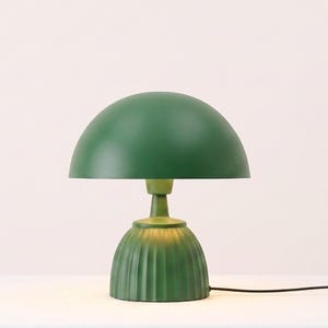 May include: A table lamp featuring a smooth, dark green dome-shaped shade resting on a matching green base with vertical fluted detailing. The lamp has a central stem connecting the shade to the base and includes a thin black power cord.