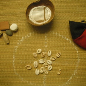 May include: A wooden bowl with a white interior, a small brown rectangular object, a white round object, a small gray rock, a brown and white shell, a red and black pouch, and a circle of white cowrie shells on a brown woven surface.