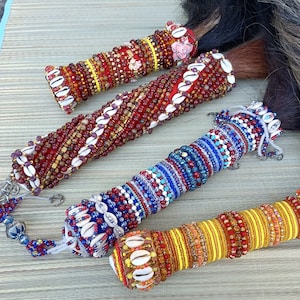 May include: Four beaded decorative wands with various color schemes. The wands are cylindrical and adorned with beads, shells, and other embellishments. The color palettes include red, blue, yellow, and white. The wands are arranged on a woven mat.