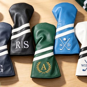 Könnte beinhalten: Fünf Golfschlägerhauben in Marineblau, Schwarz, Dunkelgrün, Blau und Weiß. Jede Haube verfügt über zwei horizontale Streifen und eine personalisierte Stickerei, darunter Initialen, ein Lorbeerkranz mit dem Buchstaben A oder gekreuzte Golfschläger.