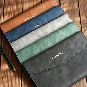 Op de afbeelding: Een stapel van vijf rechthoekige documentmappen in bruin, blauw, lichtgrijs, groen en donkergrijs, elk voorzien van een klepsluiting en een gepersonaliseerde naam of initialen in goud- of zilverkleurige letters: MICHAEL, P.R., JAMES, S.K. en ROBERT.