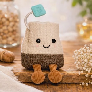 Puede incluir: Un personaje caprichoso de bolsita de té de peluche con una cara sonriente y pies de pana. La bolsita de té tiene un cuerpo beige claro, una parte inferior marrón y una etiqueta turquesa. Se encuentra sobre una superficie de madera.