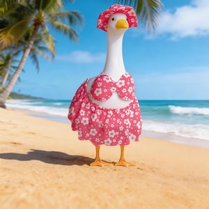 May include: A white goose figurine wearing a pink floral print bikini and matching sun hat, standing on a sandy beach with palm trees and a blue sky in the background. The goose has a yellow beak and orange feet.