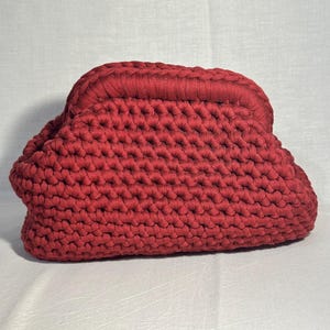 May include: A handmade, red crochet clutch bag. The bag features a textured, dense stitch pattern and a rounded shape. The top of the bag has a thicker, ribbed detail. The bag is made of a soft, flexible material.