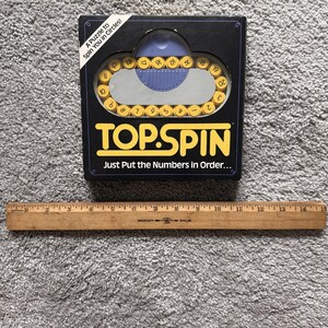 May include: A Top Spin puzzle game in a black box with yellow numbered tiles and a blue sliding mechanism. The box features the text A Puzzle to Spin You in Circles! and Just Put the Numbers in Order. A wooden ruler shows the box is 8 inches wide.