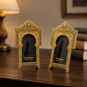 Puede incluir: Dos marcos dorados ornamentados con aberturas arqueadas. Los marcos tienen detalles intrincados y el texto "Prestigiarte OURO 24 K" es visible. Se exhiben sobre una superficie de madera oscura.