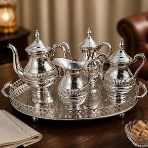 Puede incluir: Un juego de té plateado con una tetera, dos tazas, una jarra de crema y un azucarero, todo sobre una bandeja plateada decorativa. El juego se exhibe sobre una mesa de madera oscura, con una servilleta de lino y un pequeño cuenco de terrones de azúcar.
