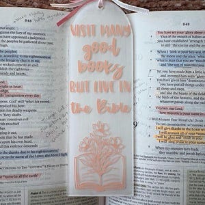Peut inclure: Marque-page en acrylique transparent avec l'inscription pêche "VISIT MANY good books BUT LIVE IN the Bible". Il présente un motif floral et un ruban rose et blanc.
