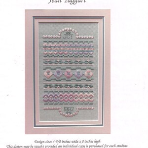 May include: A framed cross-stitch sampler by Jean Taggart featuring horizontal bands of floral and geometric patterns in shades of pink, purple, and green on a gray fabric. The text reads Design size: 4-5/8 inches wide x 8 inches high.