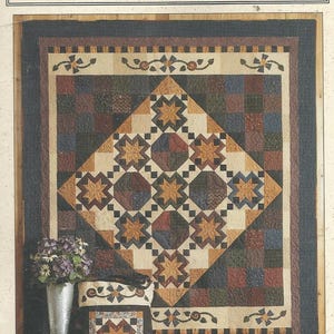 May include: A quilt pattern titled "Prairie Stars" with a large central diamond design featuring star motifs in shades of brown, beige, and blue. The quilt is accompanied by a matching tote bag and a small square item, all with similar star patterns. A vase of flowers sits to the left.