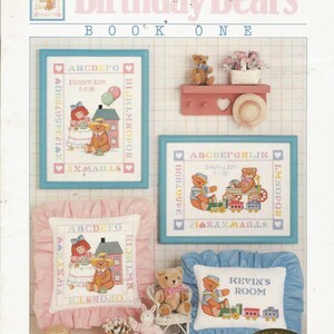May include: A book cover titled "Birthday Bears Book One" with cross-stitch designs. The designs include framed samplers, pillows, and a shelf with a teddy bear and a hat. The color palette is soft pastels, with text and illustrations.