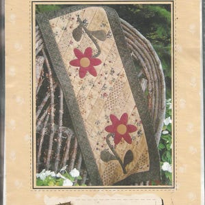 May include: A sewing pattern titled Bloomin' Runner Quilt as you go GE-117 by G.E. Designs. The cover features a table runner with a diagonal quilted pattern, floral fabric strips, and two large red flower appliqués with yellow centers and green stems.