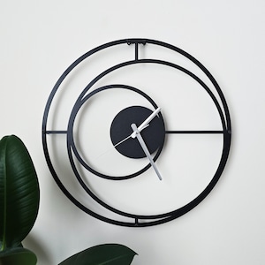 May include: A modern black metal wall clock with a unique circular design. The clock features a black circular face with silver hands and a minimalist aesthetic. The clock's outer frame has a diameter of approximately 18 inches.