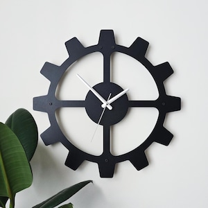 Può includere: Un orologio da parete nero a forma di ingranaggio con lancette bianche. L'orologio ha un centro circolare e un design a dente di ingranaggio attorno al perimetro. L'orologio è montato su una parete bianca. Il diametro dell'orologio è di circa 61 cm.