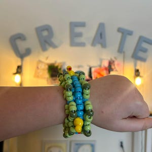 May include: A beaded bracelet in shades of green, blue, and yellow. The bracelet is made of small, round beads and features a yellow charm. The word "CREATE" is visible in the background.