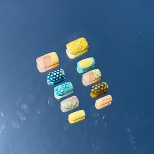 May include: A collection of ten colorful, artificial nails with various designs. The nails feature patterns like polka dots, floral, and geometric shapes in shades of blue, yellow, pink, and brown. Each nail is approximately 0.75 inches long.