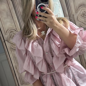 May include: A light pink blouse with ruffled detailing and a pearl belt. The blouse features a button-down front and intricate cut-out patterns along the seams. The sleeves are long and also have ruffled detailing. The blouse is made of a lightweight fabric.