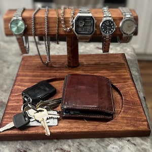 May include: A wooden valet tray with a dark stain, holding a brown leather wallet, car keys, and several watches and necklaces. The watches have silver and black bands and faces. The necklaces are silver. The tray is designed to organize everyday carry items.