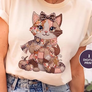 Puede incluir: Una camiseta color crema con un gráfico de un gato estilo patchwork que lleva un lazo y una bufanda con estampado de leopardo, sosteniendo una calabaza. El diseño del gato incorpora varios patrones marrones, naranjas y florales. Una insignia circular dice 1 DIGITAL PRINT.
