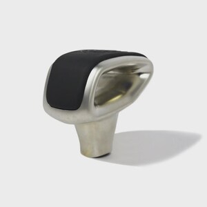 Könnte beinhalten: Ein moderner Schaltknauf mit schwarzem Oberteil und silberfarbenem Metallkörper. Der Knauf hat ein abgerundetes, ergonomisches Design und ist vor einem schlichten weißen Hintergrund platziert.