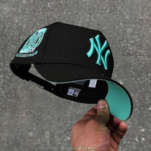May include: Black baseball cap with a teal brim and teal embroidered "NY" logo. The cap features a teal patch on the side with text. The underside of the brim is teal. The cap is held up by a hand.