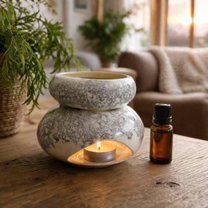 Op de afbeelding: Een tweedelige keramische etherische olie diffuser met een gespikkeld grijs en wit ontwerp. Een brandende theelichtkaars zit in de basis en een kleine amberkleurige glazen fles met een zwarte dop staat ernaast. De diffuser staat op een houten tafel.