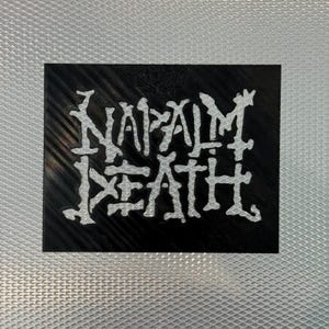 May include: A black square with the white text "NAPALM DEATH" in a stylized font. The background is a textured silver surface. The image is a close-up shot.