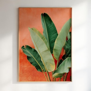 May include: A vertical rectangular wall art print featuring large, vibrant green banana leaves set against a textured, solid coral-orange background. The leaves display detailed veining and natural variations in shade, creating a bold contrast.