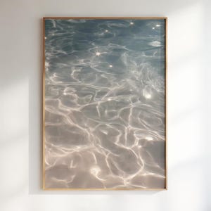 Puede incluir: Una impresión artística enmarcada que presenta una imagen abstracta de agua. La superficie del agua es una mezcla de azul claro y blanco, con reflejos brillantes. El marco es de color marrón claro y la obra se exhibe contra una pared blanca.