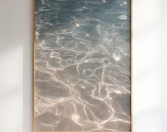 May include: A framed art print featuring an abstract image of water. The water's surface is a mix of light blue and white, with shimmering highlights. The frame is a light brown color, and the artwork is displayed against a white wall.