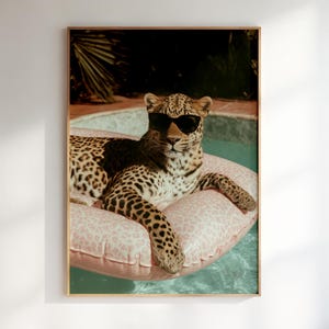 May include: A leopard with black spots and tan fur wearing black sunglasses, resting on a light pink inflatable pool float with a subtle leopard print pattern, positioned in a swimming pool with clear blue water and a palm tree in the background.