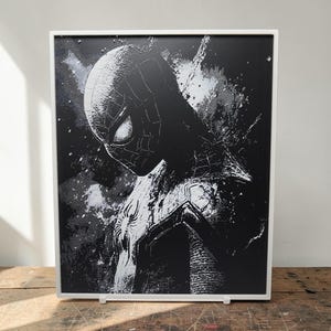 Puede incluir: Obra de arte enmarcada en blanco y negro que representa a Spider-Man. La imagen muestra al superhéroe en una pose dinámica, con una estética texturizada, casi arenosa. El marco blanco proporciona un contraste limpio con la obra oscura.
