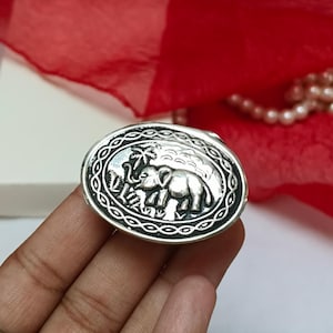 May include: Oval silver-colored ring with an embossed design of two elephants and foliage. The ring has a decorative border and is set against a red background. The ring is held by a hand.