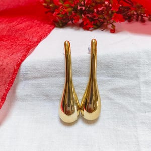 May include: A pair of gold-colored teardrop earrings. The earrings have a smooth, polished finish and a long, slender post. The background includes a red fabric and a sprig of red flowers.