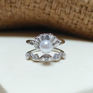 Könnte beinhalten: Ein silberfarbener Ring mit einer zentralen, muschelförmigen Fassung, die eine einzelne weiße Perle hält. Die Ringschiene ist zweigeteilt, wobei der untere Teil ein wellenförmiges Design mit fünf kleinen, klaren, rund geschliffenen Steinen aufweist.