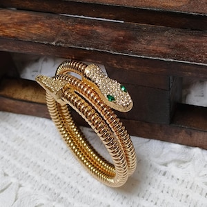 Puede incluir: Un brazalete en espiral con forma de serpiente de color dorado con un cuerpo de metal texturizado y flexible. La cabeza de la serpiente está adornada con pequeños cristales claros y dos piedras verdes de talla marquesa para los ojos.