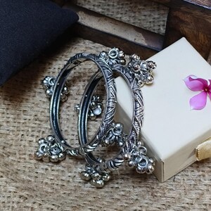 May include: Two circular silver-toned metal bangles featuring intricate etched geometric patterns and multiple small dangling spherical charms attached along the outer edges. The bangles have a dark oxidised finish in the recessed areas.