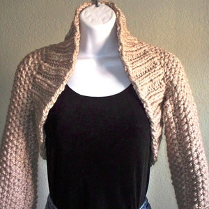 May include: A beige knitted shrug with long sleeves and a wide, open neckline. The shrug is made of a soft, textured yarn and has a casual, relaxed fit.