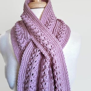 Pode incluir: Uma echarpe de malha macia cor malva com um padrão rendado delicado. A echarpe está drapeada sobre um manequim branco.