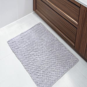 May include: A rectangular, light gray bath mat with a woven pattern. The mat is placed on a white tiled floor next to a dark brown wooden cabinet. The mat's texture is visible, suggesting a soft, absorbent material.