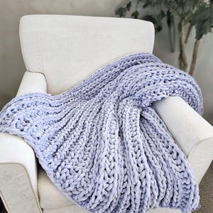May include: A light blue, chunky knit blanket draped over a white armchair. The blanket's thick, soft texture is visible, with large, looped stitches creating a cozy appearance. The chair is set against a neutral background, suggesting a comfortable living space.