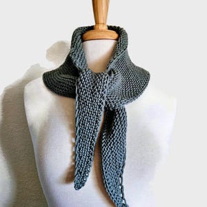 May include: A grey knitted cowl scarf displayed on a mannequin. The scarf has a wide collar that narrows into two long, pointed ends. The knitted texture is visible, and the scarf is a solid, muted grey colour.