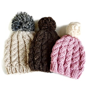 Puede incluir: Tres gorros de punto con pompones. Los gorros son beige, marrón y rosa. Los pompones son grises, marrones y blancos.