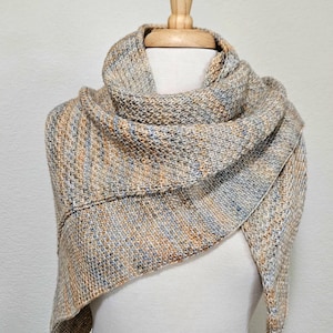 May include: A triangular knitted shawl featuring a variegated colour palette of soft tan, light blue, and cream tones. The accessory is crafted with a textured garter stitch pattern, displaying a consistent blend of these neutral and cool hues throughout.