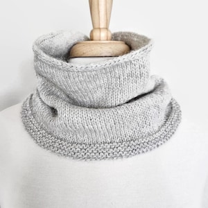 May include: A light gray knitted cowl neck warmer. The cowl is displayed on a wooden mannequin head. The neck warmer has a ribbed bottom edge and a rolled top edge. The yarn appears to be a soft, textured blend.