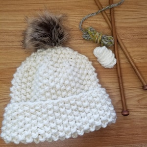 May include: A white knitted beanie with a brown faux fur pom pom. The beanie is sitting on a wooden surface with two wooden knitting needles and a ball of yarn.