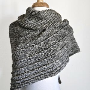 以下が含まれることがあります： グレーと白のニットのショールで、リブの模様があります。ショールは白いマネキンにかけられています。