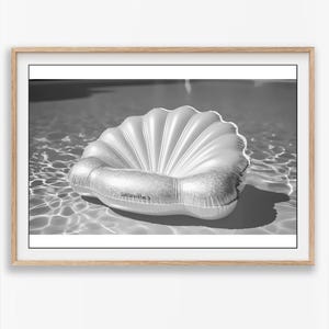 May include: A black and white photograph of an inflatable seashell pool float resting on the water surface. The float features a ribbed, fan-like design with a smooth, rounded base. The text "Sunnylife" is printed on the front of the float.