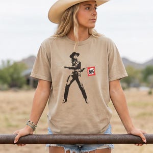 May include: Tan short-sleeved t-shirt featuring a black and white graphic of a cowgirl holding a gun and a red flag with the word "BANG". The cowgirl is wearing a hat and boots. The shirt is a relaxed fit.