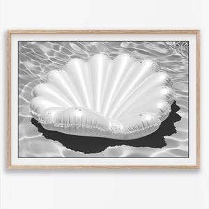 May include: A black and white framed wall art print featuring a large, inflatable seashell pool float resting on the surface of rippling water. The shell has a distinct ribbed, fan-like structure with a glossy, reflective surface and a dark shadow.